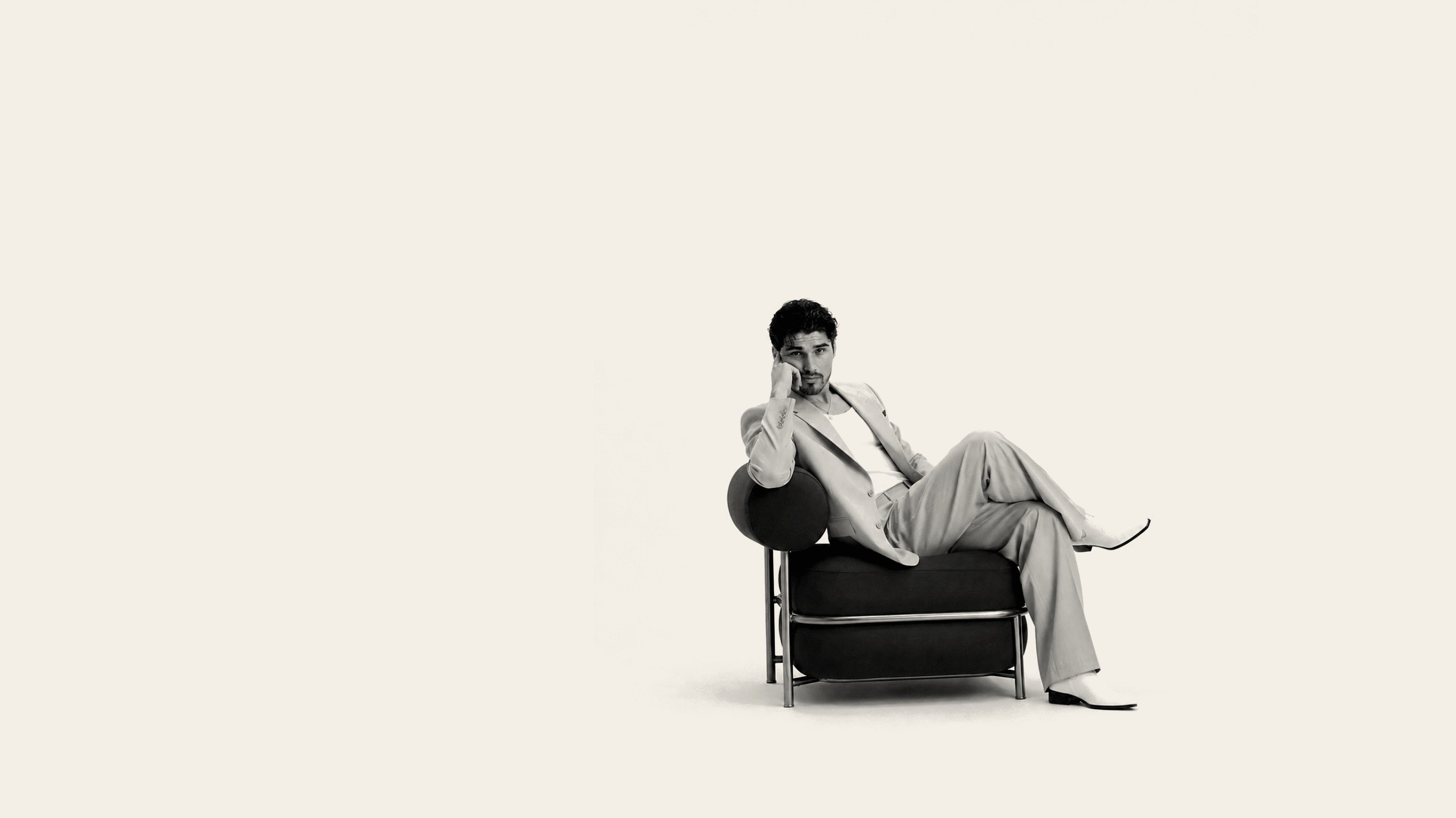 Sebastian Soriani seated in a black armchair in a light suit, one leg crossed, chin resting on his hand. Black-and-white studio portrait.